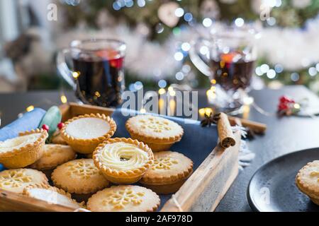 Composition de confortable chaud anglais fête traditionnelle pâtisserie mince pies en plateau en bois avec arrière-plan flou de vin chaud des boissons, une guirlande de feux Banque D'Images