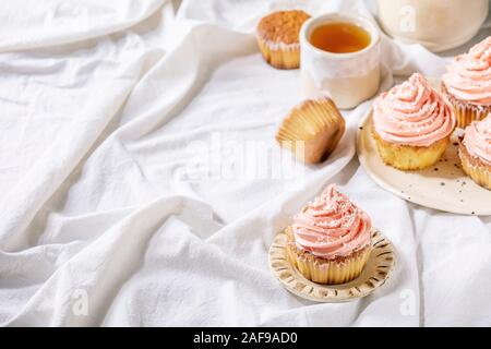 Petits gâteaux faits maison avec crème au beurre rose et flocons de noix de coco servi avec théière en céramique, tasse de thé blanc sur nappe pliée. Banque D'Images