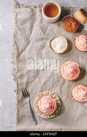 Petits gâteaux faits maison avec crème au beurre rose et flocons de noix de coco servi avec une tasse de thé en céramique sur toile gris en arrière-plan. Mise à plat, de l'espace Banque D'Images