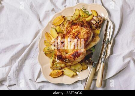 Poulet rôti entier avec des pommes de terre, l'ail et des citrons sur grand plat de céramique vintage avec fourchette et couteau sur toile tableau blanc comme arrière-plan. Mise à plat, Banque D'Images