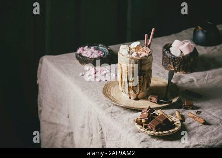 Chocolat chaud ou de cacao avec de la guimauve servi en tasse avec soucoupe en céramique, des bâtons de cannelle, chocolat, sucre rose et pot de crème sur l'onglet gris Banque D'Images