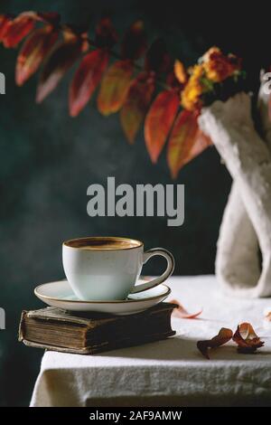 Tasse de café noir espresso debout sur nappe blanche dans une pièce sombre avec les feuilles d'automne et des fleurs dans vase d'argile, de vieux livres. Banque D'Images