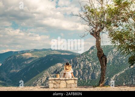 Jeune femme en robe a la mode assis sur becnch en pierre en face de Delfi paysage de montagne à Delphes, Grèce Banque D'Images