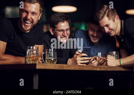 Contenu drôle sur smartphone. Trois amis se reposant dans la pub avec une bière dans les mains. Avoir conversation Banque D'Images