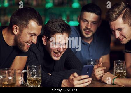 Contenu drôle sur smartphone. Trois amis se reposant dans la pub avec une bière dans les mains. Avoir conversation Banque D'Images