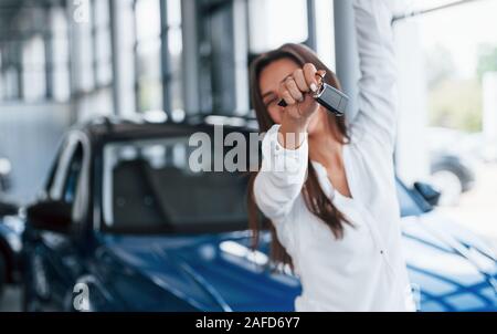 Heureux avec sa nouvelle voiture. Détient les clés dans la main. Jeune femme en blanc vêtements officiels se place en avant de l'intérieur automobile bleu Banque D'Images