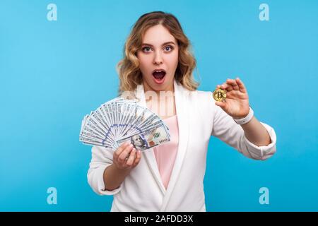 Cryptocurrency. Portrait de surprise charmante femme avec les cheveux ondulés en veste blanche holding golden physique et bitcoin, dollars de publicité btc cr Banque D'Images