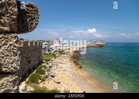 Le long de la côte dans le port de Mandraki Rhodes Ville avec plage de gravier, l'eau turquoise cristalline, vieux moulins, Kastell Agios Nikolaos et de la lumière Banque D'Images