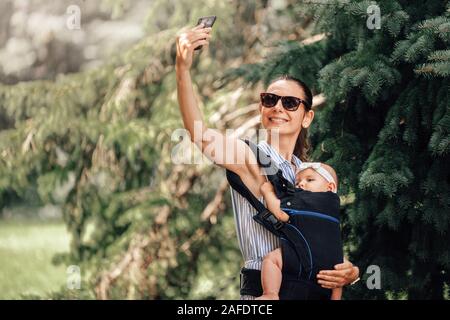 Mère avec son enfant dans un porte-bébé en tenant avec blogger ou smartphone selfies Banque D'Images