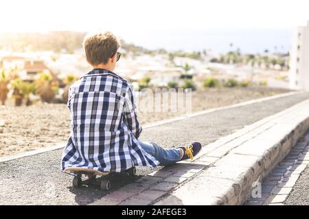 Jeune garçon assis sur son skateboard bénéficiant d'une très belle soirée à l'extérieur. Des ados à la mode d'avoir du plaisir avec la raie, la lumière du soleil et panorama maritime Banque D'Images