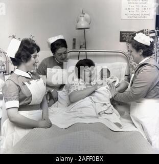 Années 1960, historique, dans une maternité, une nouvelle mère couchée dans son lit d'hôpital tenant son nouveau bébé soutenu par des infirmières rassemblées autour d'elle, Angleterre, Royaume-Uni. Un panneau au-dessus du lit indique les heures de la maternité où il était permis de fumer. Banque D'Images