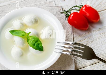 Mini boules de mozzarella au basilic dans un bol et deux tomates cerises rouges sur fond blanc table en bois rustique. Une cuisine méditerranéenne d'appui. Banque D'Images