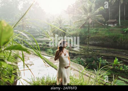 Jeune femme enceinte en robe blanche avec vue sur les rizières en terrasses de Bali dans la lumière du soleil du matin. Harmonie avec la nature. Concept de la grossesse. Banque D'Images