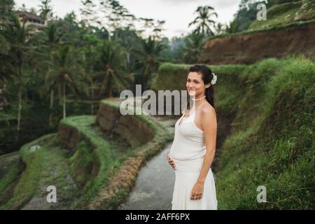 Jeune femme enceinte en robe blanche avec vue sur les rizières en terrasses de Bali dans la lumière du soleil du matin. Harmonie avec la nature. Concept de la grossesse. Banque D'Images