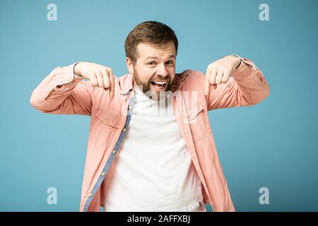 Homme barbu a l'air heureux et son index vers le bas, tout sourire et en regardant la caméra. Isolé sur un fond bleu Banque D'Images