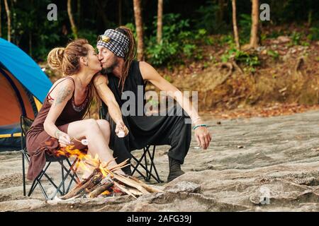 Kissing couple élégant assis sur des chaises pliantes par le feu et les guimauves de friture Banque D'Images