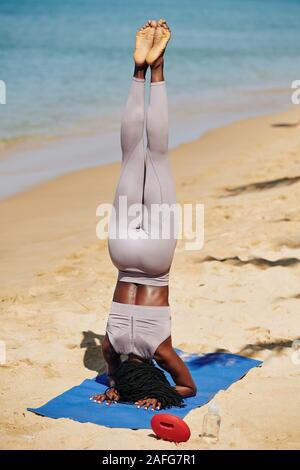 Jeune femme noire sportive l'exécution de la tête sur un tapis de yoga sur la plage, vue de l'arrière Banque D'Images