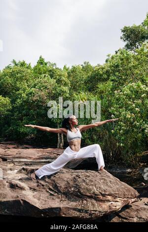 Belle jeune femme noire debout sur un gros rocher dans la forêt à distance en position deux guerrier et bénéficiant d'une séance de yoga Banque D'Images