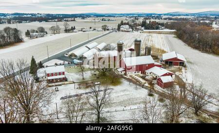 Ferme traditionnelle américaine avec bâtiments rouge derrière les arbres après la première chute de neige en automne, vue aérienne du paysage rural avec petite rivière Banque D'Images