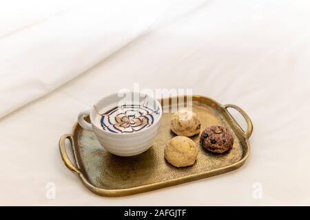 Cappuccino avec fantaisie design chocolat sur le haut avec trois différents biscuits cuits sur le plateau d'or dans chambre d'hôtel sur bed Banque D'Images