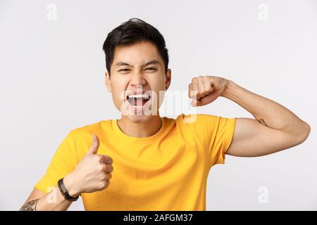 Gros plan audacieux et confiant beau asian man in yellow t-shirt, montrant le muscle et pouce, criant a encouragé et excité, sentir Banque D'Images