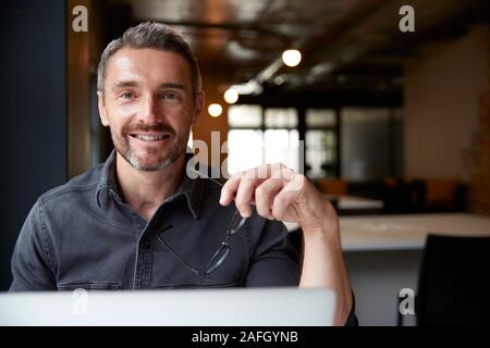 Homme de race blanche d'âge moyen creative sitting at desk in office holding glasses, souriant à la caméra, Close up Banque D'Images
