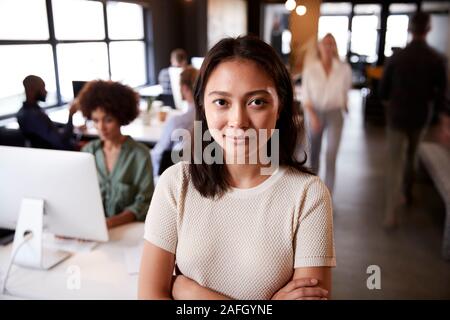 Femme Asiatique millénaire creative debout dans un bureau temporaire occupé, souriant à l'appareil photo Banque D'Images