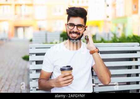 Jeune homme assis sur un banc, parler au téléphone, en regardant la caméra. Bel homme en portant des lunettes en blanc shirt élégant. Il worker smiling, pos Banque D'Images