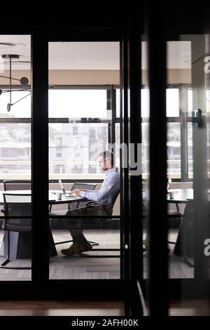 Homme blanc d'âge moyen qui travaille seul dans une salle de réunion, vu à travers la paroi de verre, vertical Banque D'Images