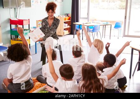 Les élèves du primaire en uniforme lever la main pour répondre à la question que de femmes enseignant lit Book Banque D'Images