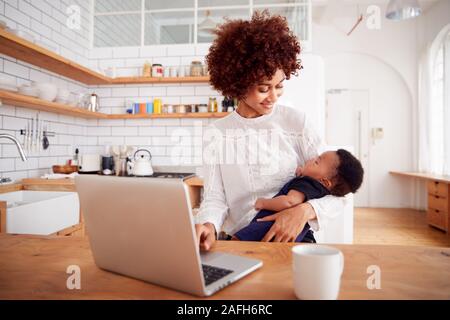 Multitâche mère détient bébé endormi Fils et travaille sur un ordinateur portable dans la cuisine