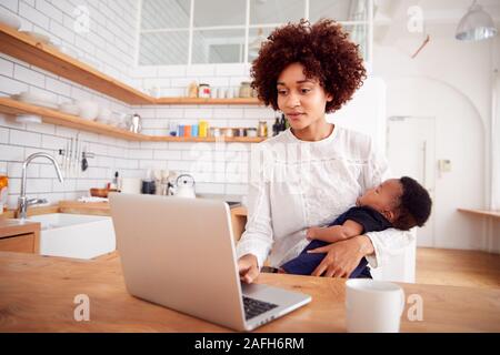 Multitâche mère détient bébé endormi Fils et travaille sur un ordinateur portable dans la cuisine