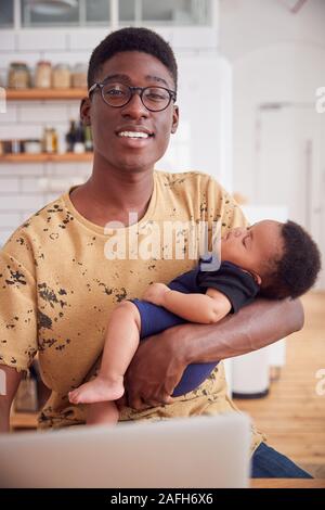 Portrait du père multitâches Holding Baby son sommeil et working on laptop computer in Kitchen Banque D'Images