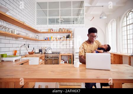 Multitâche père détient bébé endormi Fils et travaille sur un ordinateur portable dans la cuisine Banque D'Images