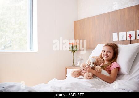 Portrait of Girl Lying in Bed In Hospital Ward Hugging Teddy Bear Banque D'Images