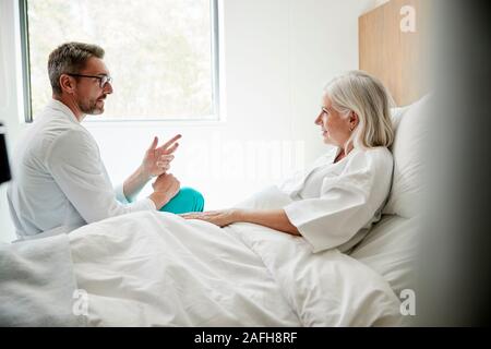 Médecin Wearing White Coat Visiter Mature Female Patient In Hospital Bed Banque D'Images