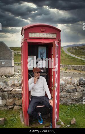 Homme assis dans une boîte de téléphone rouge traditionnelle éloignée ayant une conversation, Isle of Harris, Outer Hebrides, Ecosse Banque D'Images