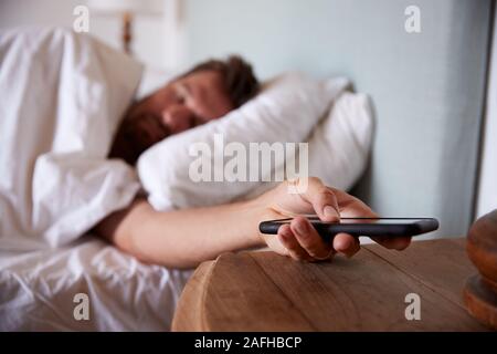 Mid adult man endormi dans son lit, tendant la main vers le smartphone sur la table de chevet à l'avant-plan Banque D'Images