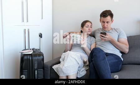 Jeune couple, homme et femme choisissez budget hôtel pour se reposer à l'aide de téléphone mobile. Femme est compter les espèces de l'argent. Ils sont assis sur le canapé de leur appartement. Banque D'Images