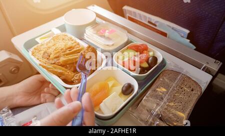 Déjeuner dans l'avion : pain, viande avec légumes et riz, des morceaux de fromage, d'abricot séché, de salades et de boissons dans la tasse, la lumière du soleil. Femme est de manger en avion pendant le vol de la nourriture servie sur plateau. Banque D'Images