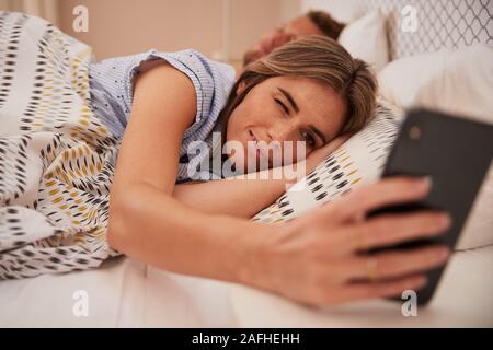 Femme blanche à moitié endormi dans bed looking at smartphone, son partenaire dormant dans background, Close up Banque D'Images