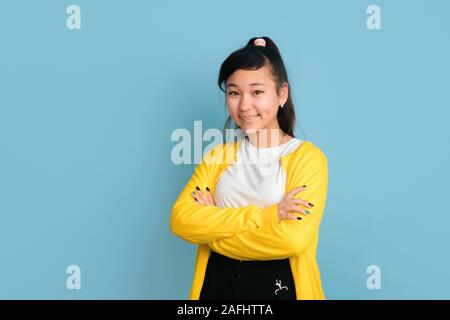 Asian teenager's portrait isolé sur fond studio bleu. Belle brunette model femme aux cheveux longs. Concept d'émotions humaines, expression du visage, ventes, ad. Posant avec les mains croisées. Banque D'Images