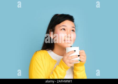 Asian teenager's portrait isolé sur fond studio bleu. Belle brunette model femme aux cheveux longs. Concept d'émotions humaines, expression du visage, ventes, ad. Boire du café ou du thé. Banque D'Images