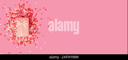 Boîte cadeau rose avec un arc dans le centre de l'image sur fond de bonbons coeur doux peu dispersées sur un fond rose. Vue de dessus. Banque D'Images