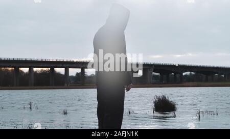 Un sombre, Moody, hiver éditer d'un capuchon transparent fantomatique figure éphémère et debout à côté d'un lac, à la recherche sur un pont de l'autoroute, avec une unité d'organisation Banque D'Images