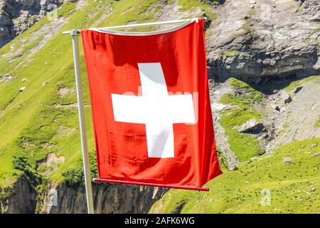 Brandissant le drapeau national de la Suisse avec paysage alpin flou en arrière-plan. Croix blanche sur la place rouge. Symbole national du pays. Drapeau suisse. Banque D'Images