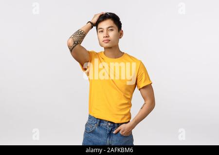 Confiant et élégant beau mec tatoué chinois en jaune t-shirt, exécuter les doigts dans les cheveux et à l'auto-assurance, l'amour lui-même et l'appareil photo Banque D'Images