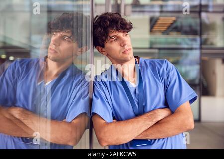 Jeune médecin homme fatigué de porter des gommages corporels Leaning Against Wall dans la construction de l'hôpital moderne Banque D'Images