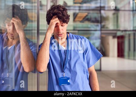 Jeune médecin homme fatigué de porter des gommages corporels Leaning Against Wall dans la construction de l'hôpital moderne Banque D'Images