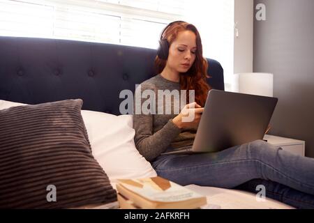 Female College Student Wearing Headphones travaille sur lit dans chambre partagée avec ordinateur portable et téléphone mobile Banque D'Images
